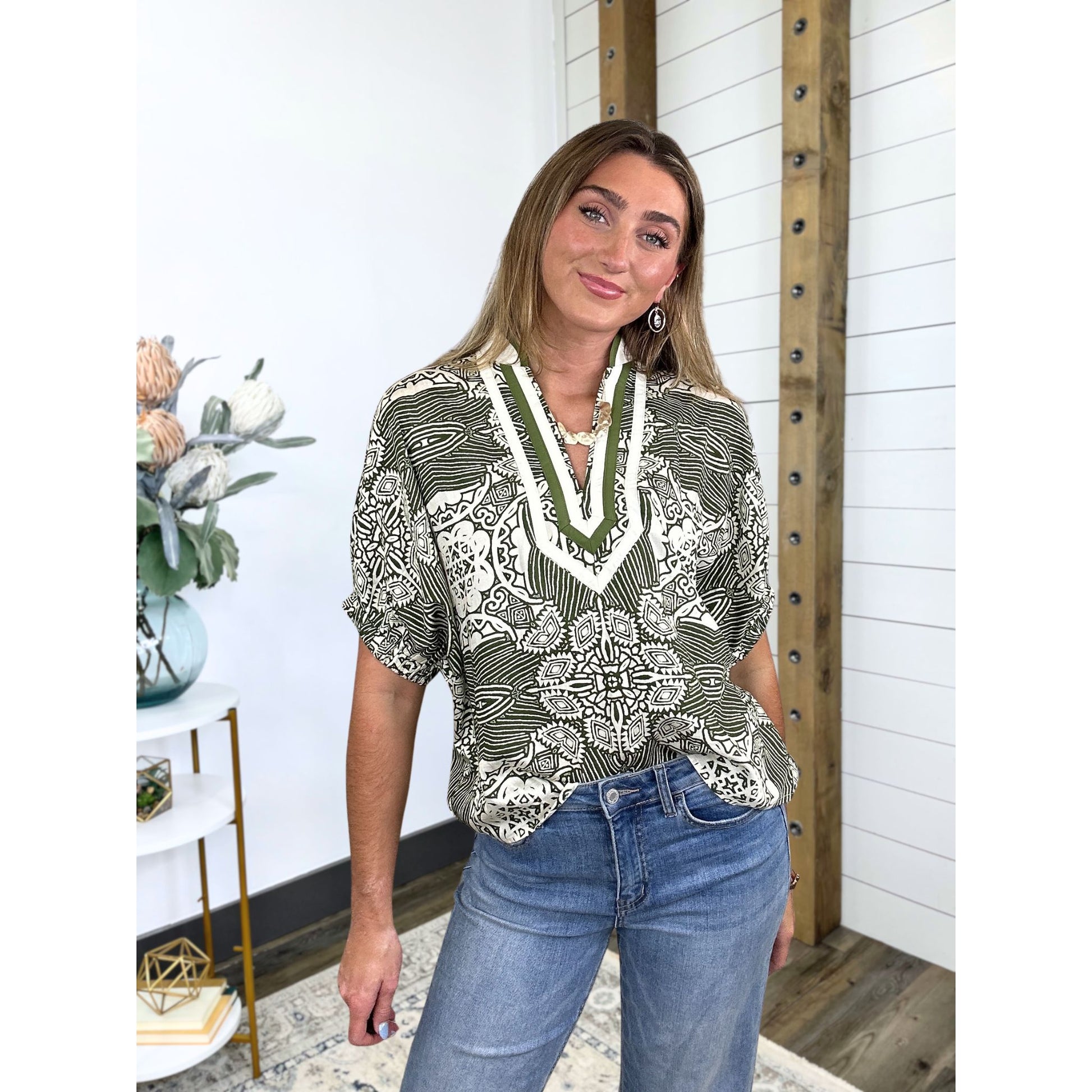 Woman wearing a patterned blouse and jeans in a room with decorative elements.