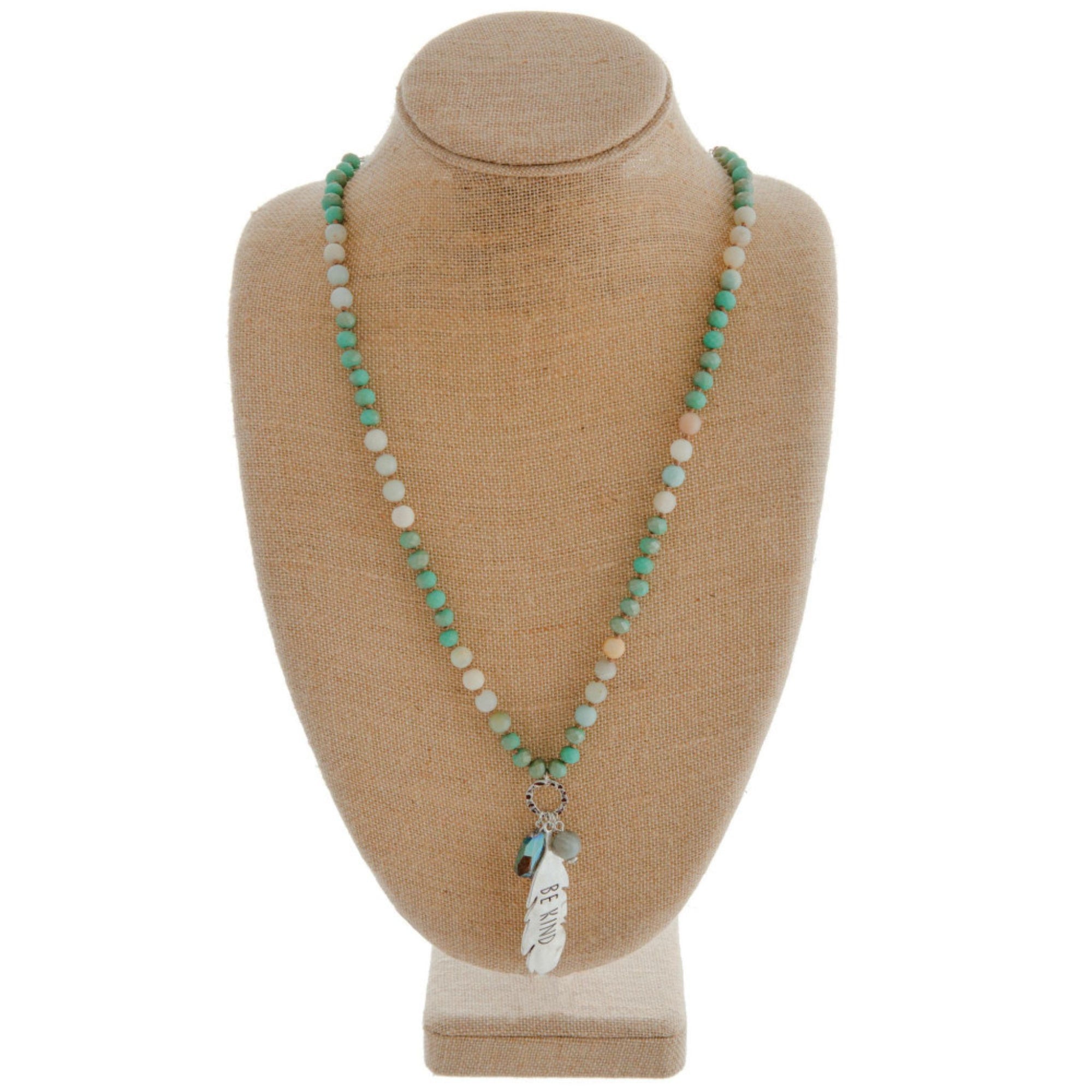 Long beaded necklace featuring a crystal accent and a metal feather shaped pendant engraved with the phrase "Be Kind."