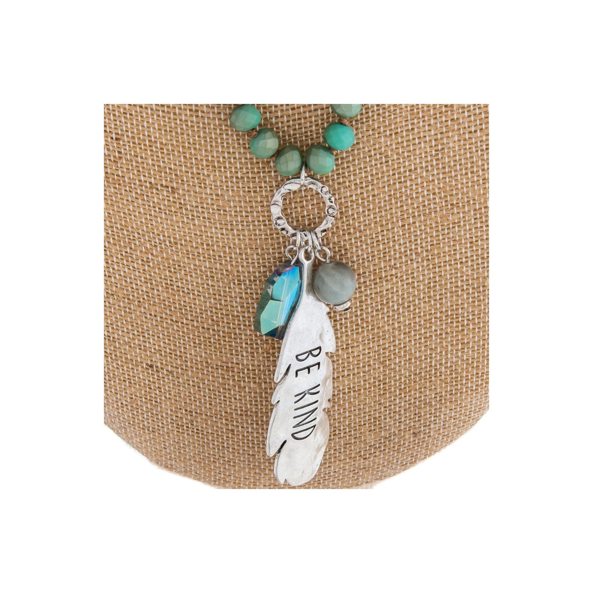 Long beaded necklace featuring a crystal accent and a metal feather shaped pendant engraved with the phrase "Be Kind."