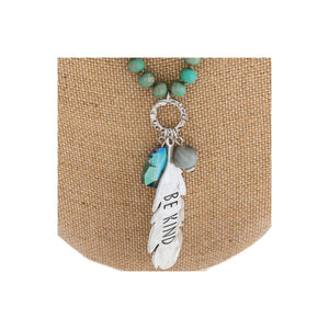 Long beaded necklace featuring a crystal accent and a metal feather shaped pendant engraved with the phrase "Be Kind."
