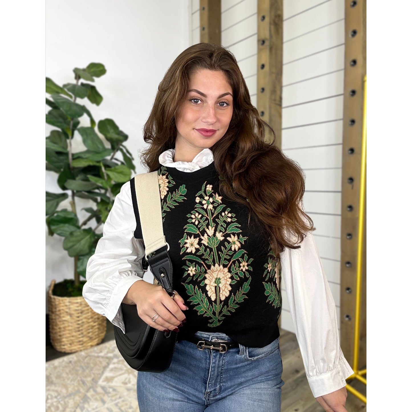 Woman wearing a black sweater with floral embroidery over a long sleeve white ruffle top, holding a black bag, standing indoors.